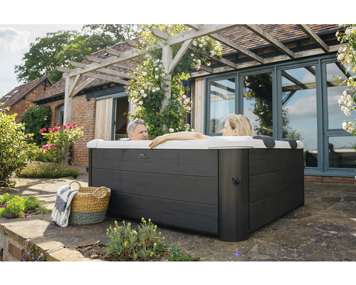 Dvě osoby relaxují ve čtvercové venkovní vířivce s tmavě šedým dřevěným obložením na terase u domu s pergolou.