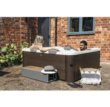 Čtvercová vířivka s dřevěným dekorem, dvě osoby relaxují ve vodě na terase před cihlovou zdí.