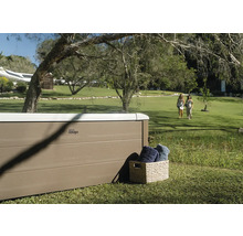 Vířivka MSpa s panely ve vzhledu dřeva ve velké zahradě s trávníkem a stromy. Vedle ní je koš s ručníky.