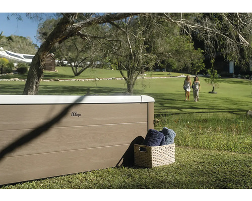 Vířivka MSpa s panely ve vzhledu dřeva ve velké zahradě s trávníkem a stromy. Vedle ní je koš s ručníky.