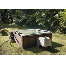 Čtvercová vířivka s obložením v dekoru dřeva a externí čerpací jednotkou, na trávníku v zahradě s palmami.