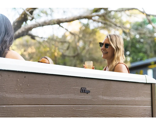 Lidé relaxují ve vířivce MSpa s exteriérem v dekoru dřeva na zahradě. Logo MSpa na bočním panelu.