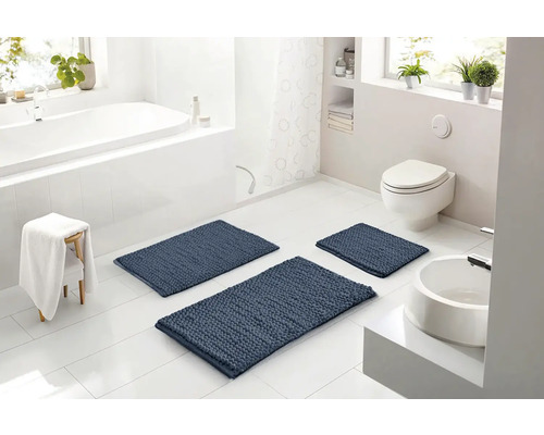 Světlá moderní koupelna se třemi modrými strukturovanými koupelnovými předložkami na bílé dlažbě, včetně vany, toalety a umyvadla.