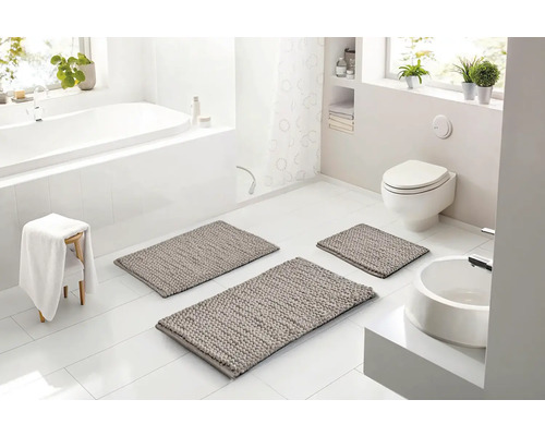Světlá, moderní koupelna se třemi šedými koupelnovými předložkami na bílé dlažbě, vanou, toaletou a umyvadlem v bílé barvě.