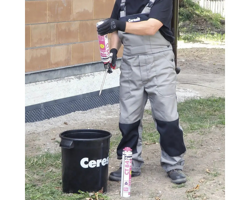 Řemeslník v pracovním oděvu drží aplikační pistoli s lepidlem Ceresit CT 84 nad kbelíkem venku.