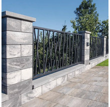 Moderní zahradní zeď z šedých tvárnic s tmavými kovovými prvky v geometrickém designu před stromy a kamennou podlahou.