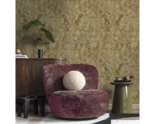 Strukturovaná tapeta s ornamentálním vzorem ve zlaté a zelené barvě v obývacím pokoji s fialovým křeslem a dřevěnou komodou.