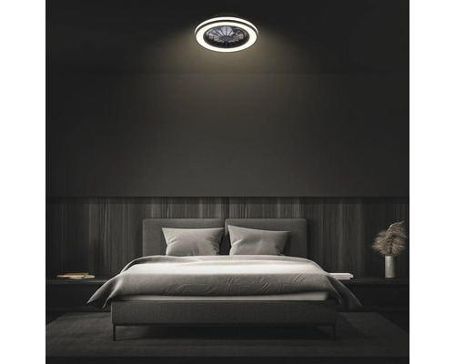 Moderní stropní ventilátor s LED osvětlením v tmavě zařízené ložnici nad šedou čalouněnou postelí.