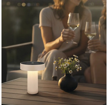 Moderní solární stolní lampa na dřevěném terasovém stole v zahradě za soumraku s květinami a lidmi v pozadí.