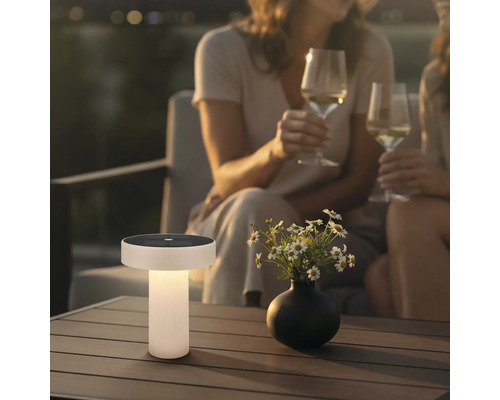 Moderní solární stolní lampa na dřevěném terasovém stole v zahradě za soumraku s květinami a lidmi v pozadí.