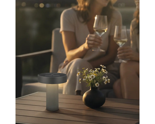 Moderní venkovní solární stolní LED lampa na dřevěném stole v útulné večerní zahradě s lidmi.