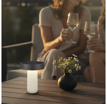 Moderní solární stolní lampa na terasovém stole vedle vázy s květinami, v pozadí dvě osoby se sklenicemi vína ve večerní atmosféře.