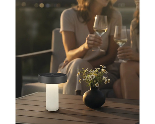 Moderní solární stolní lampa na terasovém stole vedle vázy s květinami, v pozadí dvě osoby se sklenicemi vína ve večerní atmosféře.