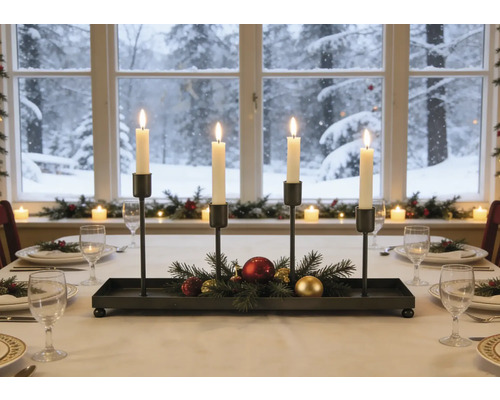 Adventní svícen z černého kovu se čtyřmi zapálenými svíčkami, jedlovými větvičkami a vánočními ozdobami na prostřeném stole.