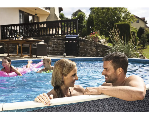 Rodina v kruhovém nadzemním bazénu na zahradě s terasou, plynovým grilem a zahradním nábytkem v pozadí za slunného dne.