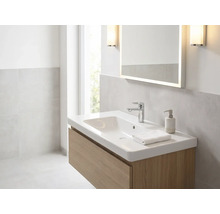 Světlá koupelna s bílým umyvadlem na dřevěné skříňce, chromovanou baterií, osvětleným zrcadlem a nástěnnými svítidly.