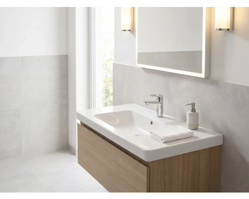 Světlá koupelna s bílým umyvadlem na dřevěné skříňce, chromovanou baterií, osvětleným zrcadlem a nástěnnými svítidly.