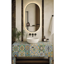 Moderní koupelna s bílým umyvadlem na barevné skříňce s dlaždicemi, podsvíceným zrcadlem, černými nástěnnými bateriemi a rostlinou.