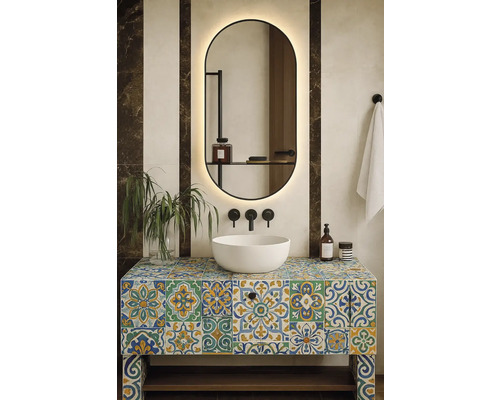 Moderní koupelna s bílým umyvadlem na barevné skříňce s dlaždicemi, podsvíceným zrcadlem, černými nástěnnými bateriemi a rostlinou.