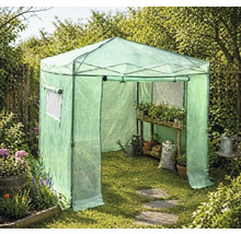 Průchozí fóliovník na zahradě s dřevěným regálem, rostlinami v květináčích, konví a pytlem se zeminou na trávníku.