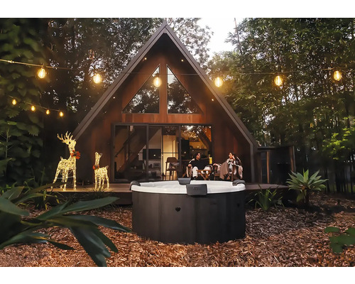 Kulatá venkovní vířivka v zahradě s moderním domem ve tvaru A, světelnými řetězy a osvětlenými dekoracemi sobů večer.