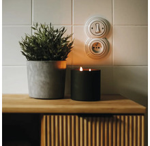 Vypínač a zásuvka v retro stylu na kachlové stěně vedle rostliny a svíčky na dřevěné komodě.