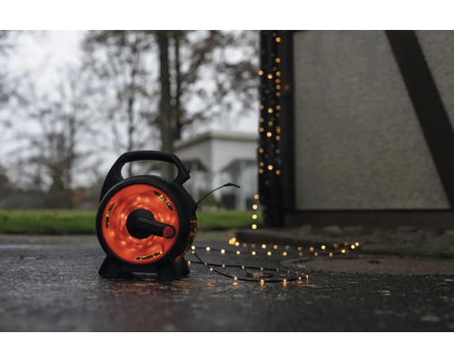 LED světelný řetěz na černém bubnu pro venkovní použití s teplým bílým světlem na mokré zemi vedle budovy.