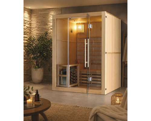 Interiérová sauna ze dřeva s proskleným čelem a dveřmi ve světlé wellness místnosti s kamennou stěnou, pokojovou rostlinou a příslušenstvím.
