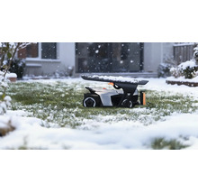 Robotická sekačka v garáži na zasněženém trávníku během sněžení před domem.