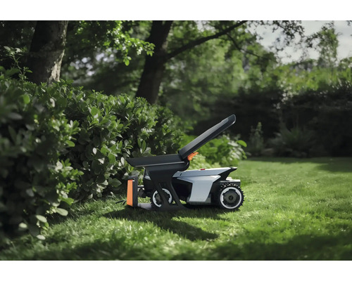 Robotická sekačka s příslušenstvím na trávníku v zahradě vedle živého plotu.