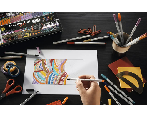 Ruka kreslí barevné vzory fixem Stabilo Creative Tips na papír, obklopená různými kreslicími potřebami a kovovým pouzdrem.