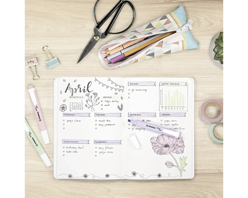 Pohled shora na světlý, uspořádaný pracovní stůl s otevřeným bullet journalem, zvýrazňovači Stabilo Swing Cool, nůžkami a papírenským zbožím.