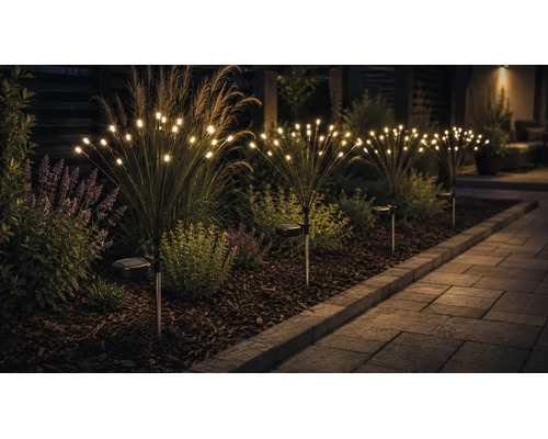 Solární zahradní svítidla v designu ohňostroje s teplými bílými LED žárovkami v záhonu vedle dlážděné zahradní cesty.