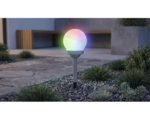 Solární zahradní kulové svítidlo s vícebarevným RGB osvětlením, na zapichovacím hrotu v záhonu s oblázky a rostlinami.