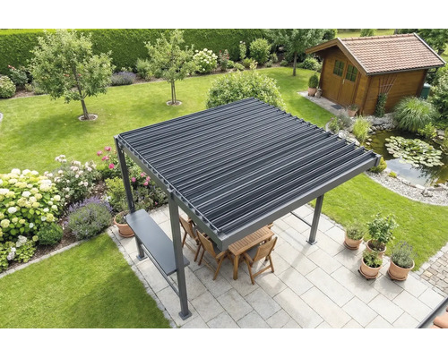 Volně stojící pergola s lamelovou střechou na světlé terase v zahradě, včetně dřevěného nábytku, přilehlého jezírka a zahradního domku v pozadí.