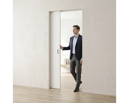 Muž v obleku otevírá bílé minimalistické posuvné dveře ve světlém, moderně zařízeném pokoji s dřevěnou podlahou.