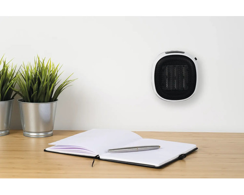 Kompaktní teplovzdušný ventilátor do zásuvky na bílé zdi nad pracovním stolem se zápisníkem, perem a dvěma pokojovými rostlinami.