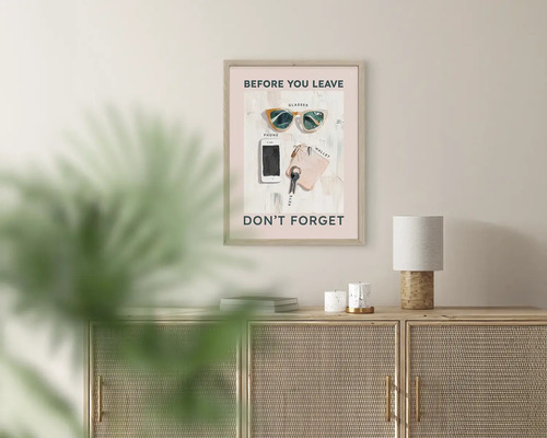 Zarámovaný plakát s textem Before you leave don't forget a ilustracemi brýlí, telefonu a klíčů nad světlou dřevěnou komodou.