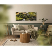 Panoramatický obraz na stěnu s motivem starého kamenného domu u potoka v moderně zařízeném, světlém obývacím pokoji s pohovkou a dřevěným stolem.