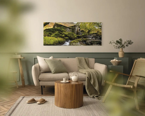 Panoramatický obraz na stěnu s motivem starého kamenného domu u potoka v moderně zařízeném, světlém obývacím pokoji s pohovkou a dřevěným stolem.