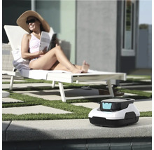 Bazénový robot na okraji bazénu, v pozadí žena na lehátku ve světlé zahradě s trávníkem a dlažbou.