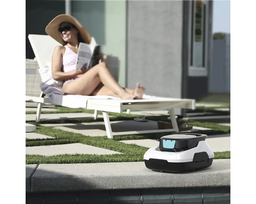 Bazénový robot na okraji bazénu, v pozadí žena na lehátku ve světlé zahradě s trávníkem a dlažbou.