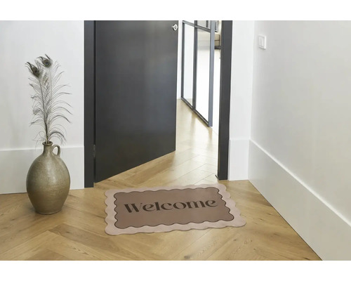 Rohožka s vlnitým okrajem a nápisem Welcome na dřevěné podlaze s rybinovým vzorem v moderní předsíni vedle tmavých dveří a vázy s pavími pery.