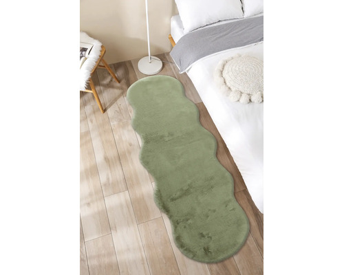 Zelený běhoun z umělé kožešiny se zvlněnými okraji ve světlé ložnici na dřevěné podlaze vedle postele.