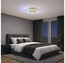 Moderní stropní ventilátor s barevným LED kruhem v ložnici s šedou čalouněnou postelí, tmavými závěsy a kobercem.