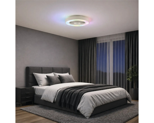 Moderní stropní ventilátor s barevným LED kruhem v ložnici s šedou čalouněnou postelí, tmavými závěsy a kobercem.