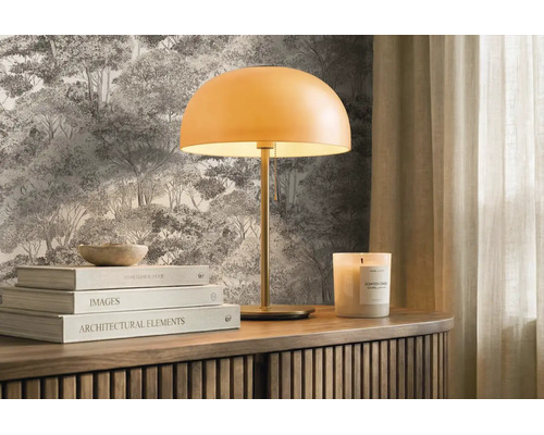 Moderní stolní lampa s kulatým stínidlem na dřevěné komodě před lesní tapetou, doplněná knihami a vonnou svíčkou. Světlé a uspořádané.