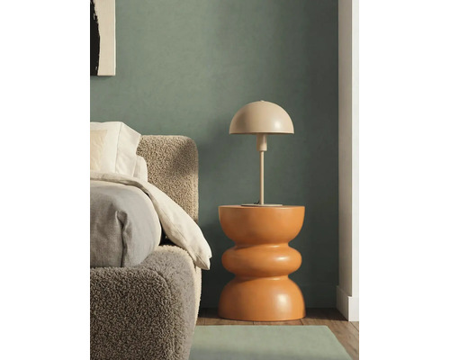 Stolní lampa ve tvaru houby na sochařském odkládacím stolku vedle postele s čelem z buklé tkaniny před tmavě zelenou stěnou.