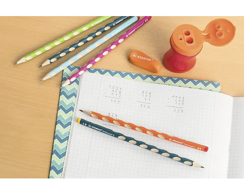 Psací stůl s ergonomickými tužkami Stabilo EASYgraph, gumou, ořezávátkem se zásobníkem a sešitem s matematickými výpočty.
