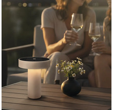 Moderní solární stolní lampa s integrovaným solárním panelem na terasové stole s vázou květin a dvěma osobami držícími sklenice vína.